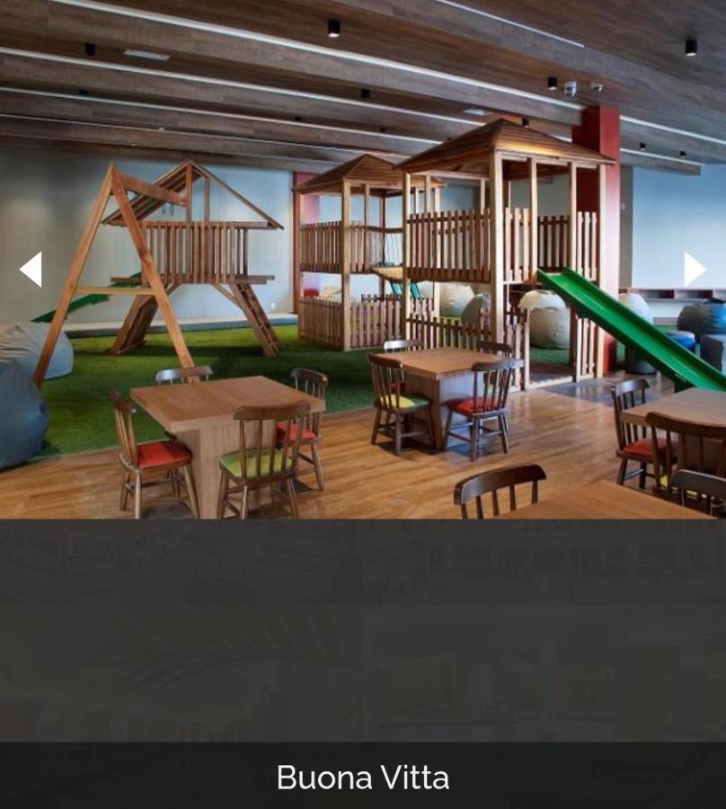 Hotel Buona Vitta Gramado by Gramado Parks Esterno foto The photo shows a play area designed for children, featuring a wooden play structure with slides and climbing areas. In the foreground, there are several small wooden tables and chairs arranged for seating. The floor is covered with green turf, resem