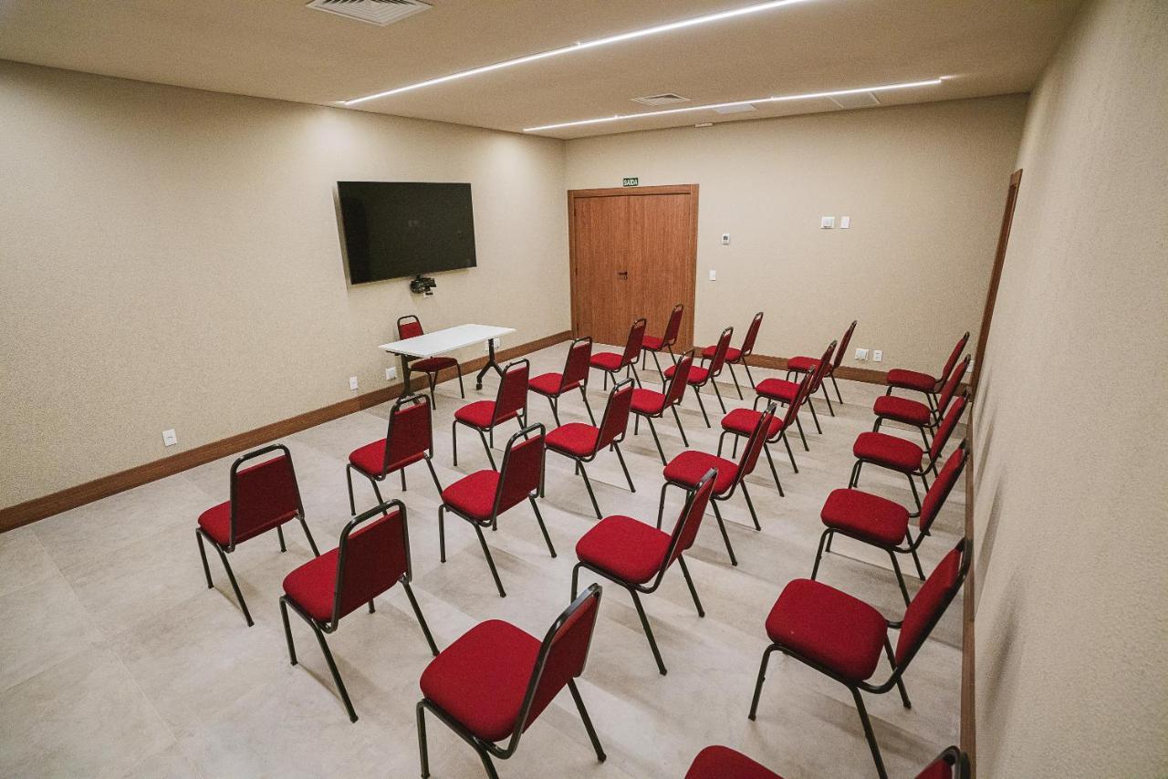 Hotel Buona Vitta Gramado by Gramado Parks Esterno foto The photo shows a conference or meeting room with a simple and modern design. There are several rows of red chairs arranged in a semi-circle facing a white table at the front. On the wall opposite the chairs, there is a flat-screen television mounted