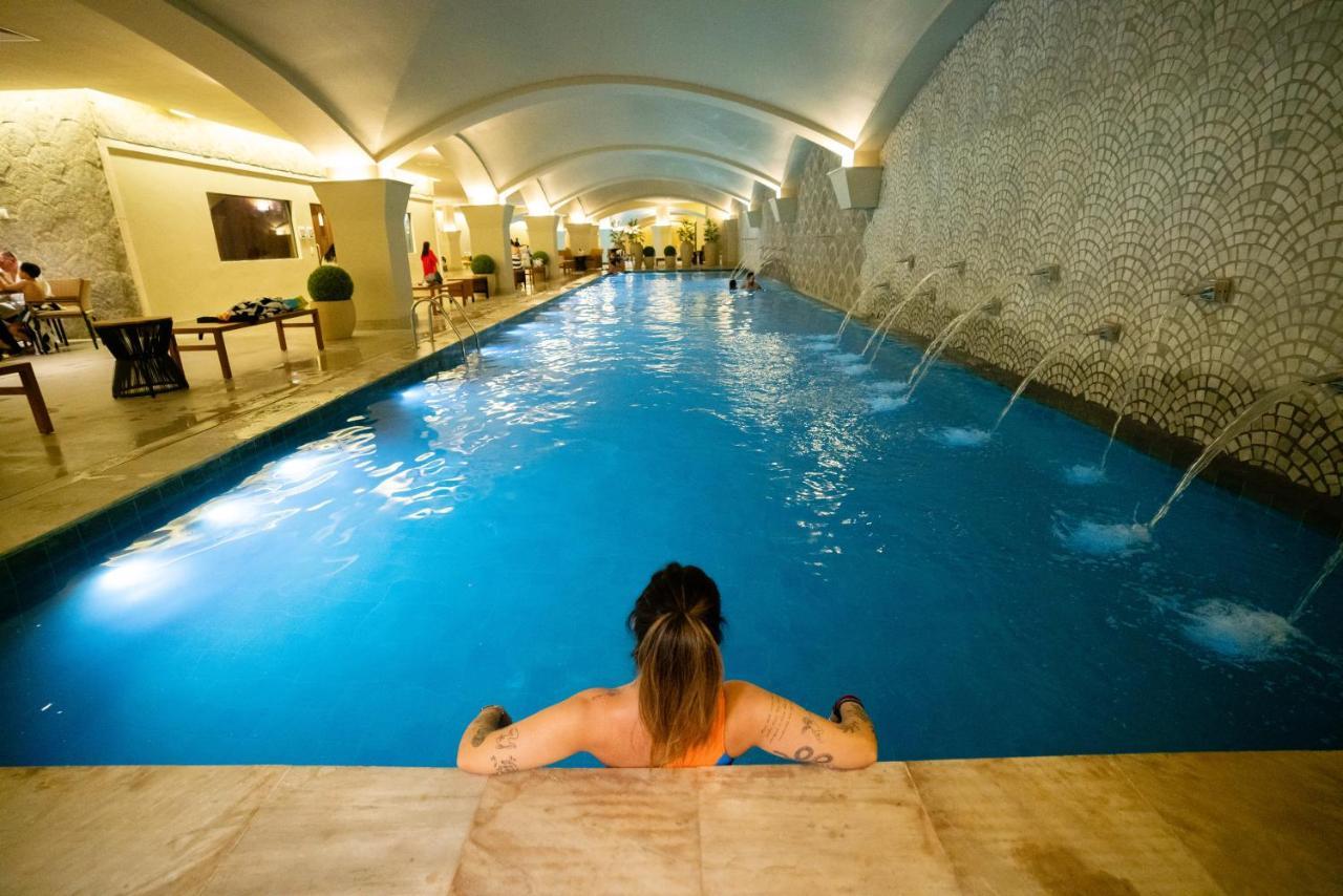 Hotel Buona Vitta Gramado by Gramado Parks Esterno foto The photo shows an indoor swimming pool with a long, narrow design. The water is a clear blue, and there are features such as fountains along one side. In the foreground, a person with long hair is sitting on the edge of the pool, looking out over th
