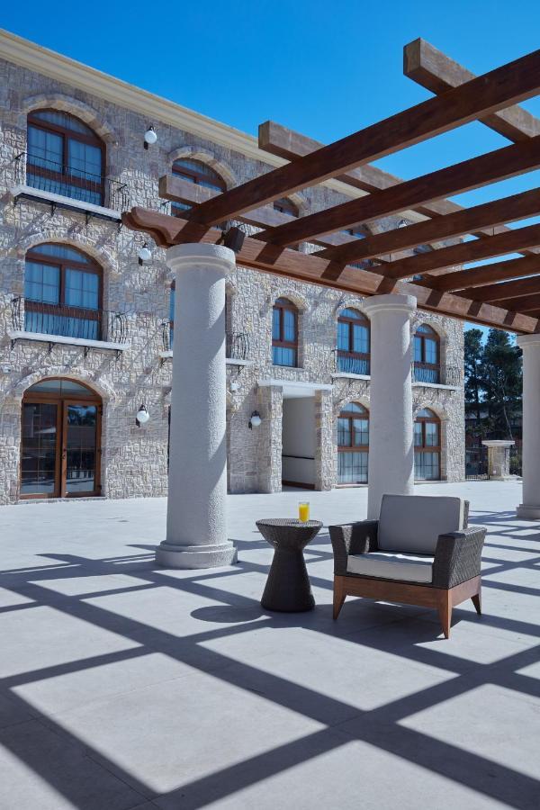 Hotel Buona Vitta Gramado by Gramado Parks Esterno foto The photo shows a well-designed outdoor space with a stone wall building in the background. There are several tall columns that support a wooden pergola overhead, creating a shaded area. In this space, there is a stylish chair and a small table besid