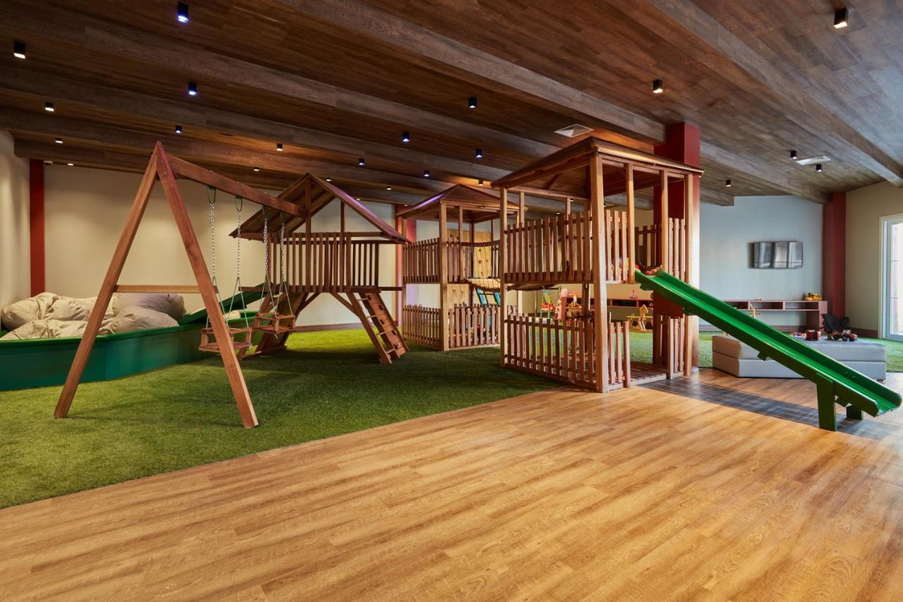 Hotel Buona Vitta Gramado by Gramado Parks Esterno foto The photo shows an indoor play area designed for children. It features a wooden play structure that includes multiple levels, slides, and a swing set. The floor is covered with wooden planks, and there is a green carpet-like area that adds a soft tou