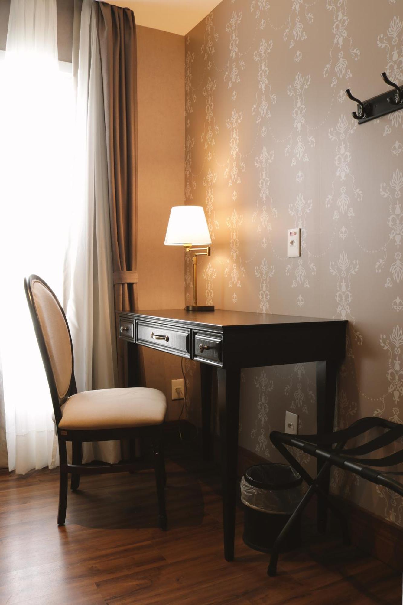 Hotel Buona Vitta Gramado by Gramado Parks Esterno foto The photo shows a small desk set against a wall with a decorative pattern. The desk is black with a few drawers and has a small lamp on it. Next to the desk, there is a chair with a light-colored cushion. Soft light is coming through a window, castin